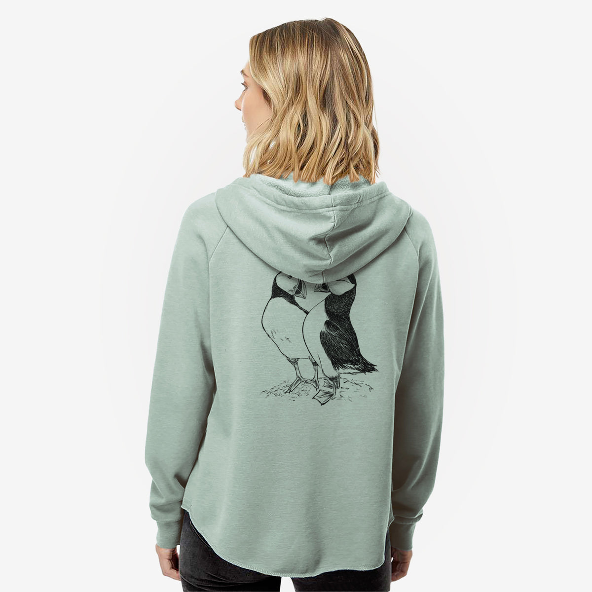 Woman wearing a sage green women's Cali Wave zip-up sweatshirt printed with a detailed black ink illustration of two Atlantic puffins standing side by side, seen from behind.