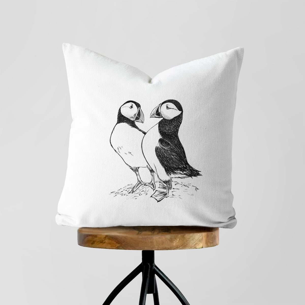 White pillow cover printed with a detailed black ink illustration of two Atlantic puffins standing side by side, on a wooden stool.