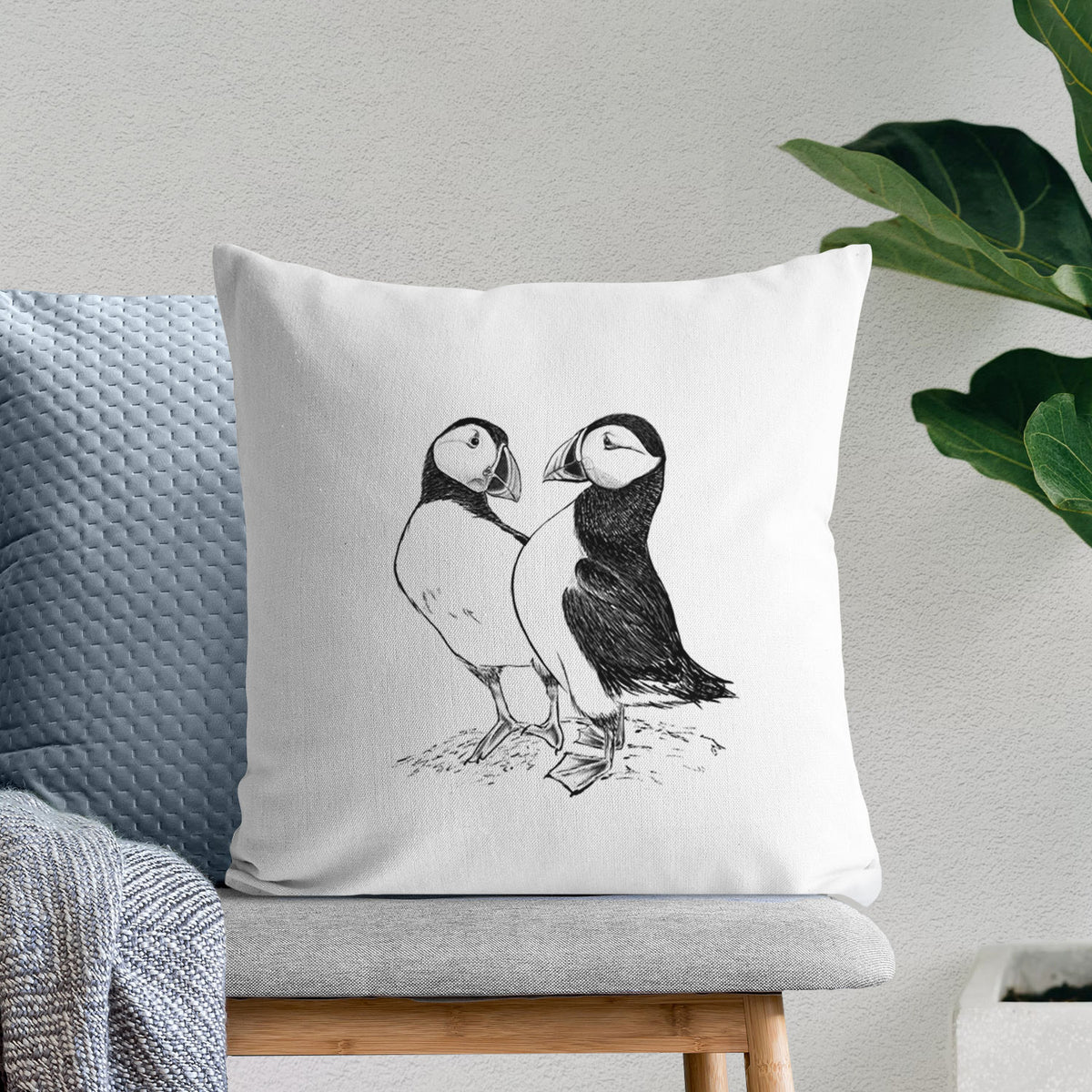 White pillow cover printed with a detailed black ink illustration of two Atlantic puffins standing side by side, on a bench beside plants.