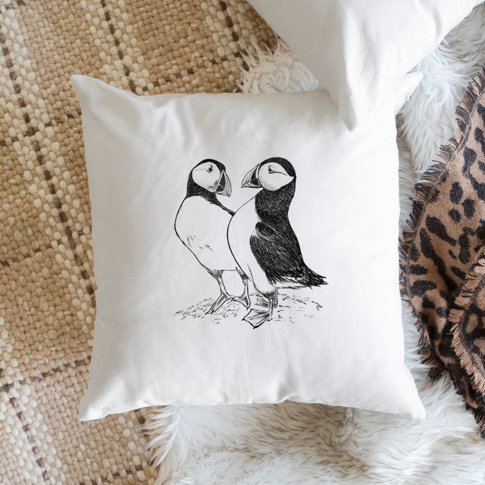 White pillow cover printed with a detailed black ink illustration of two Atlantic puffins standing side by side, on a bench beside plants.