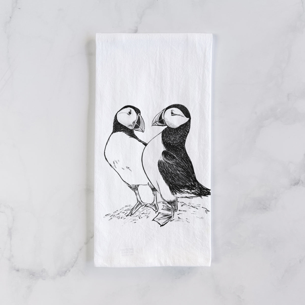 White cotton tea towel laid flat on marble, printed with a detailed black ink illustration of two Atlantic puffins standing side by side.