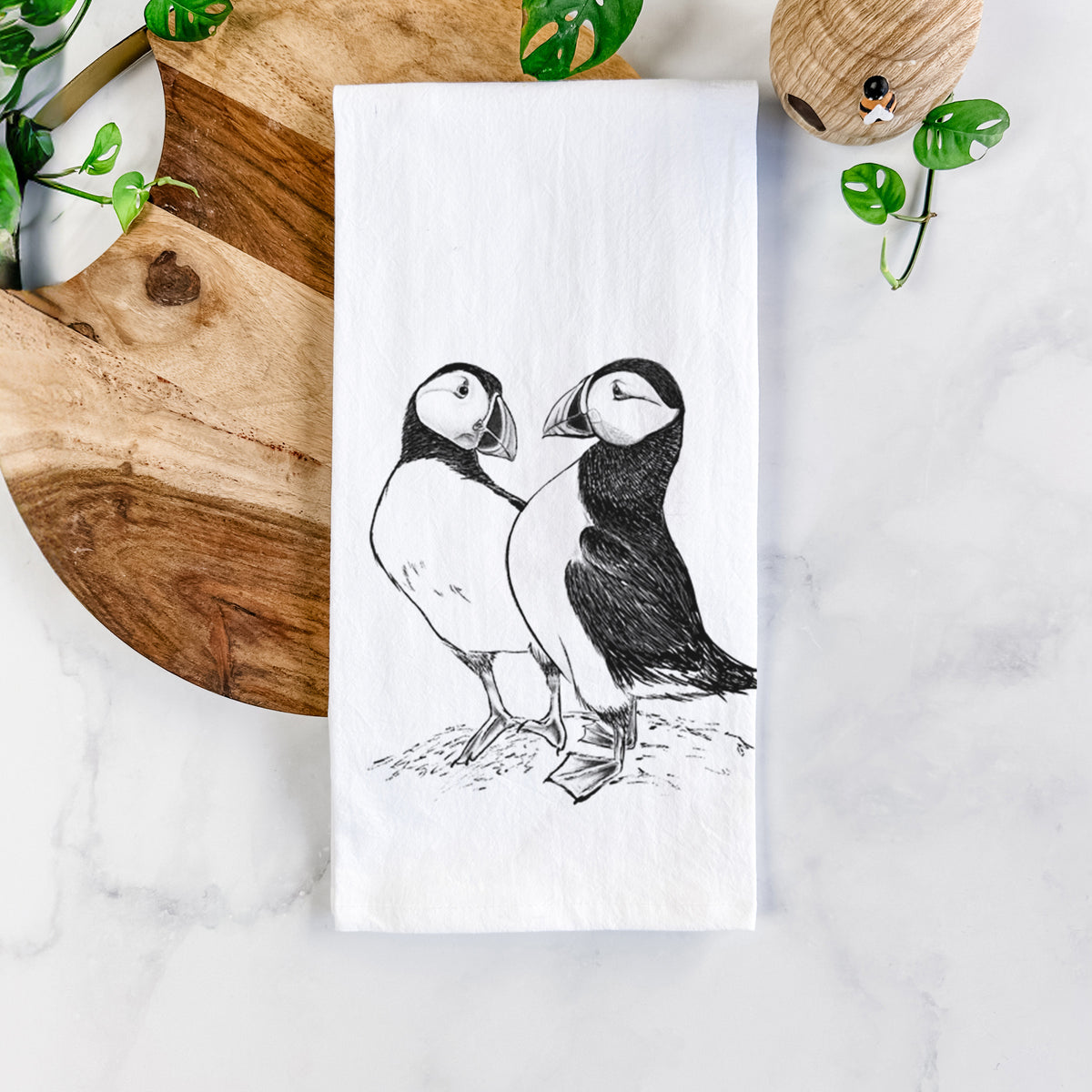 White cotton tea towel printed with a detailed black ink illustration of two Atlantic puffins standing side by side, on a marble counter beside a round wooden cutting board and greenery.