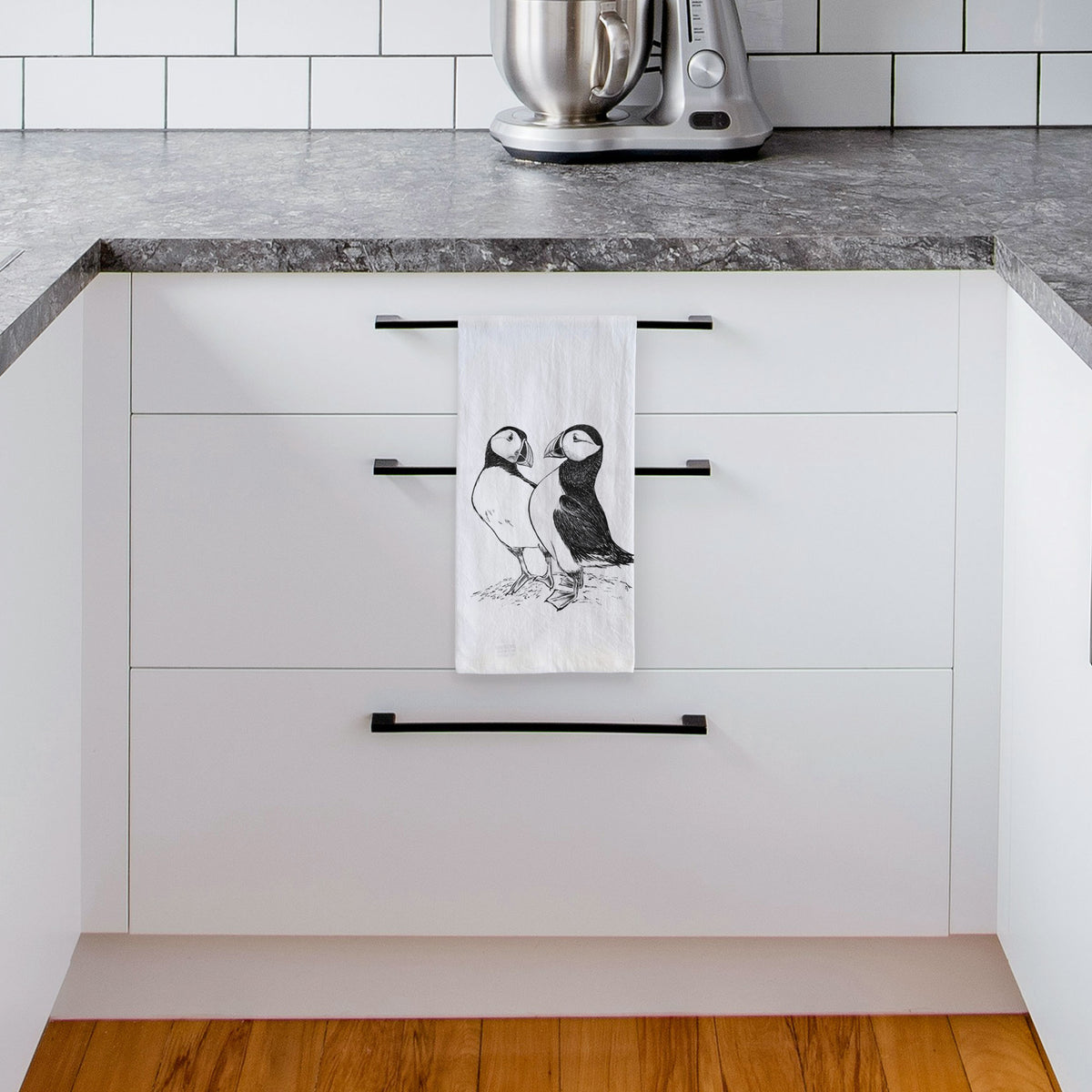 White tea towel printed with a detailed black ink illustration of two Atlantic puffins standing side by side, hanging from a drawer handle in a bright white kitchen.