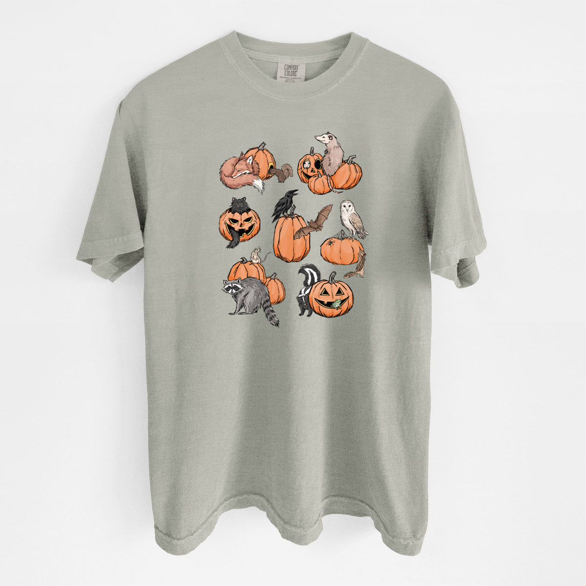 Sandstone beige Comfort Colors t-shirt laid flat, printed with orange carved jack-o'-lanterns with black ink opossums, raccoons, owls and skunks peeking out.