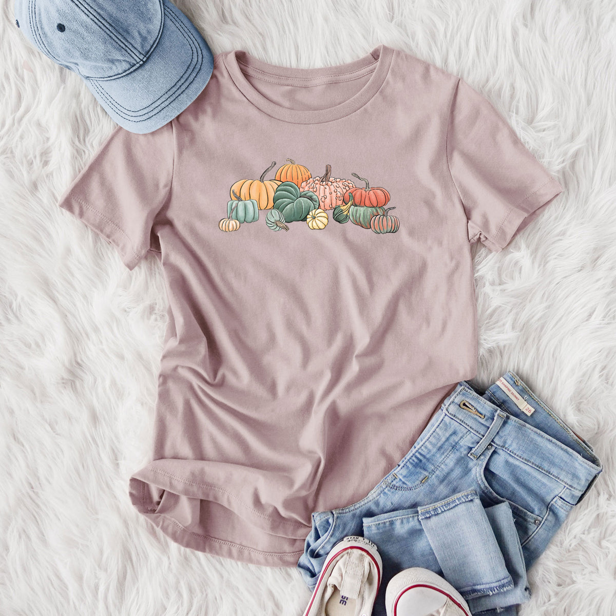 Women's relaxed fit cotton t-shirt printed with a full-color illustration of a pile of pumpkins and gourds in orange, green and white, styled flat on a white faux fur rug with jeans.