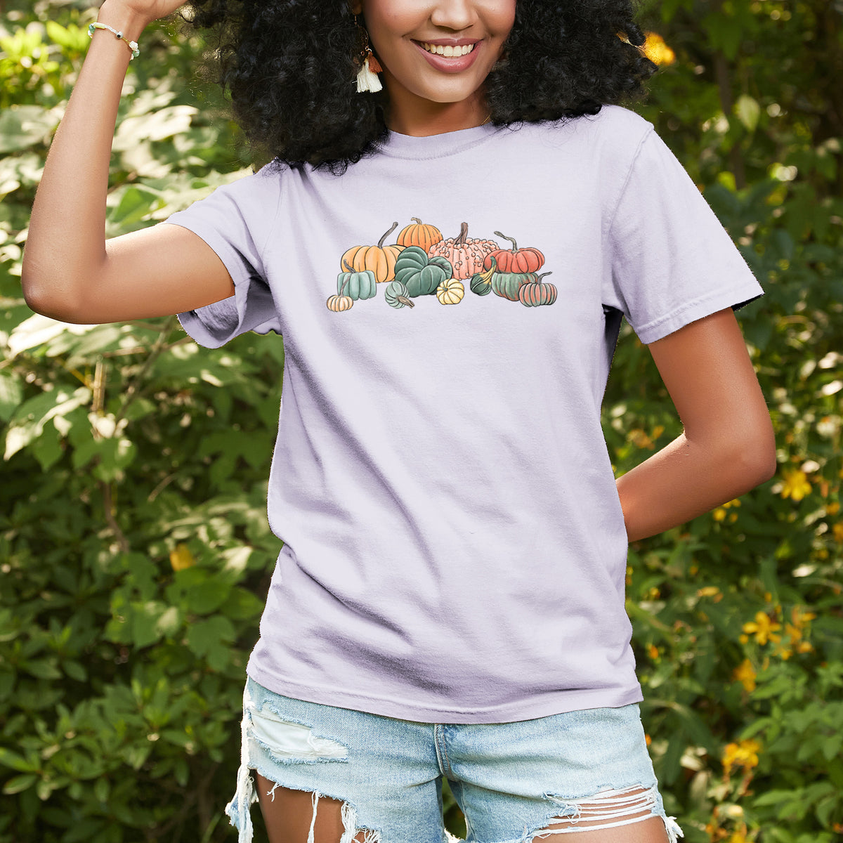 Woman wearing a Comfort Colors t-shirt printed with a full-color illustration of a pile of pumpkins and gourds in orange, green and white, outdoors.