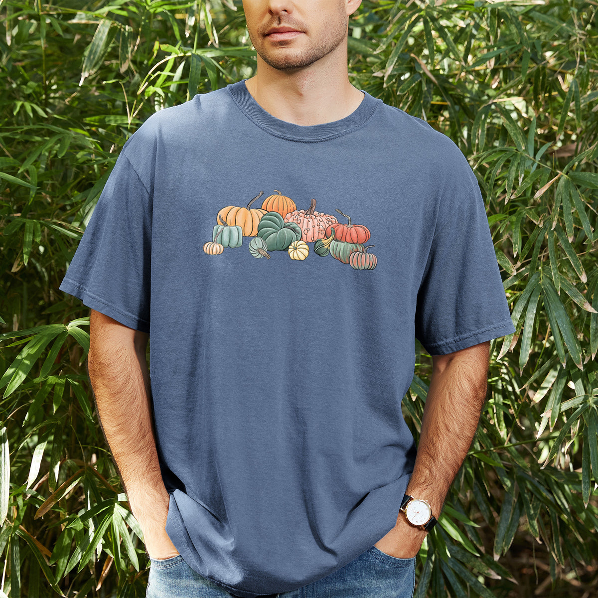 Man wearing a Comfort Colors t-shirt printed with a full-color illustration of a pile of pumpkins and gourds in orange, green and white, outdoors.