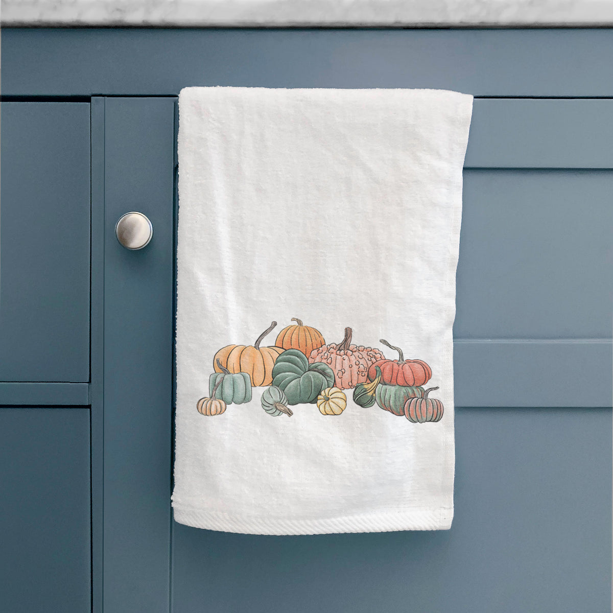White hand towel printed with a full-color illustration of a pile of pumpkins and gourds in orange, green and white, hanging on a cabinet door.