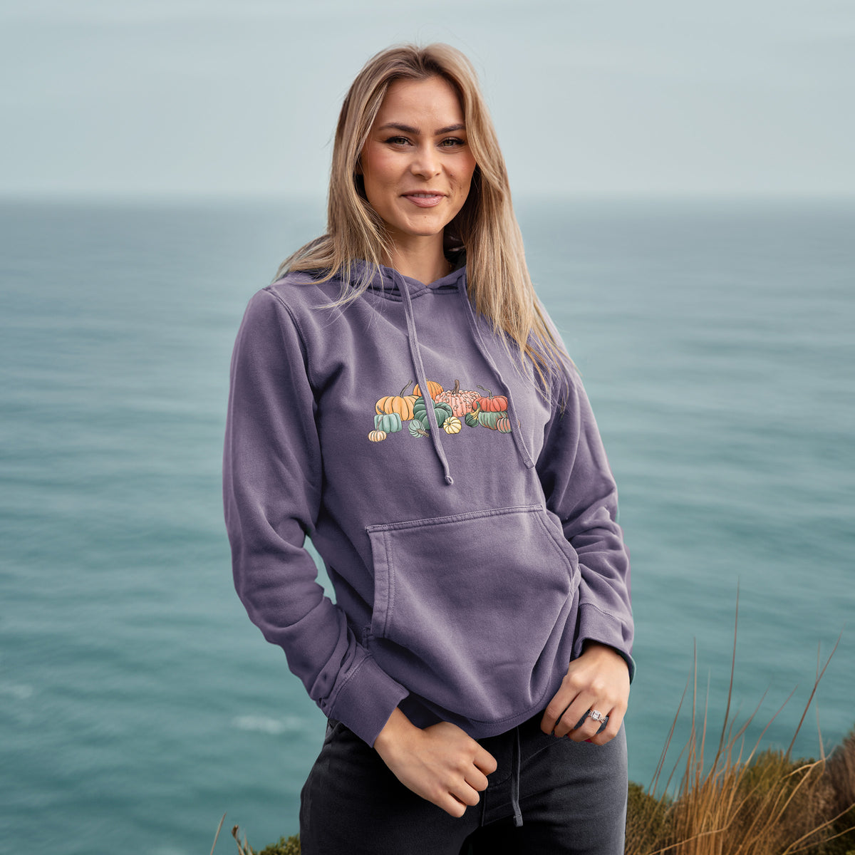 Woman wearing a plum purple pigment dyed hoodie printed with a full-color illustration of a pile of pumpkins and gourds in orange, green and white.