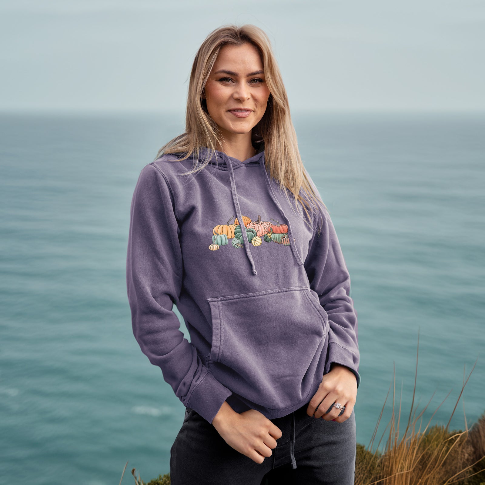 Plum purple pigment dyed hoodie laid flat, printed with a full-color illustration of a pile of pumpkins and gourds in orange, green and white.