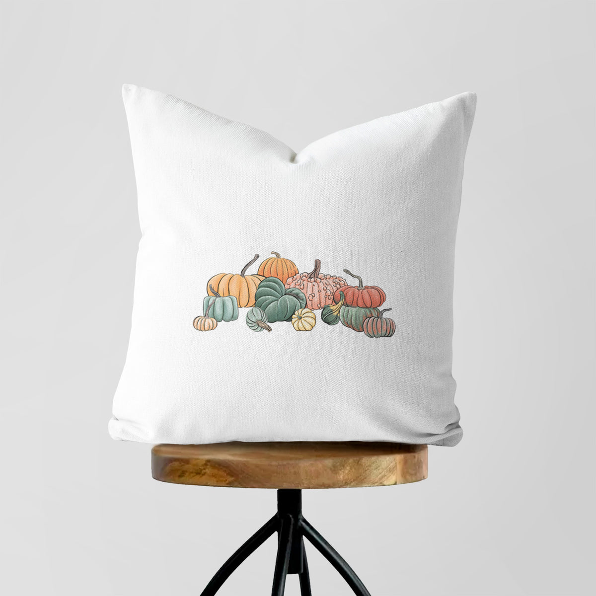 White pillow cover printed with a full-color illustration of a pile of pumpkins and gourds in orange, green and white, on a wooden stool.