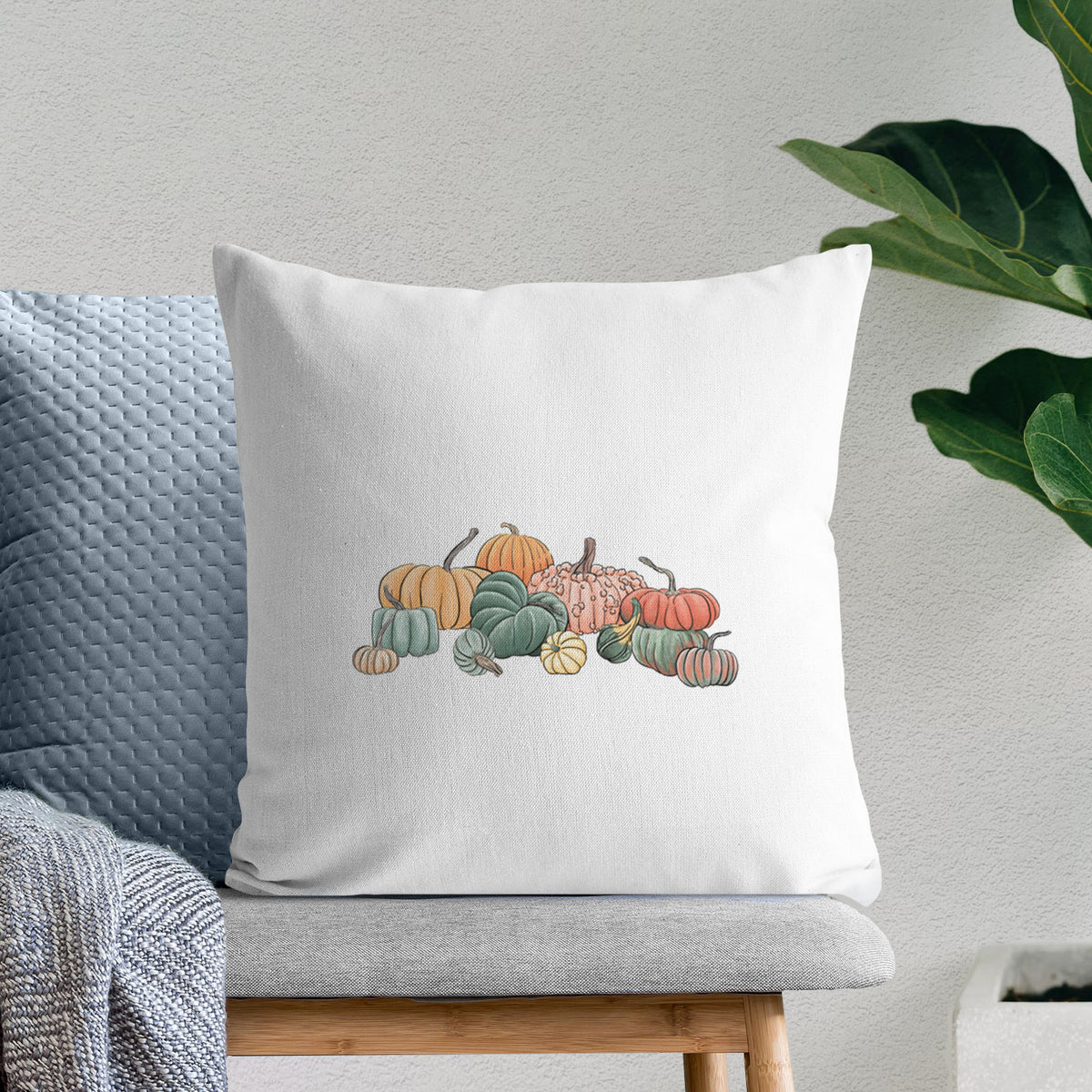 White pillow cover printed with a full-color illustration of a pile of pumpkins and gourds in orange, green and white, on a bench beside plants.