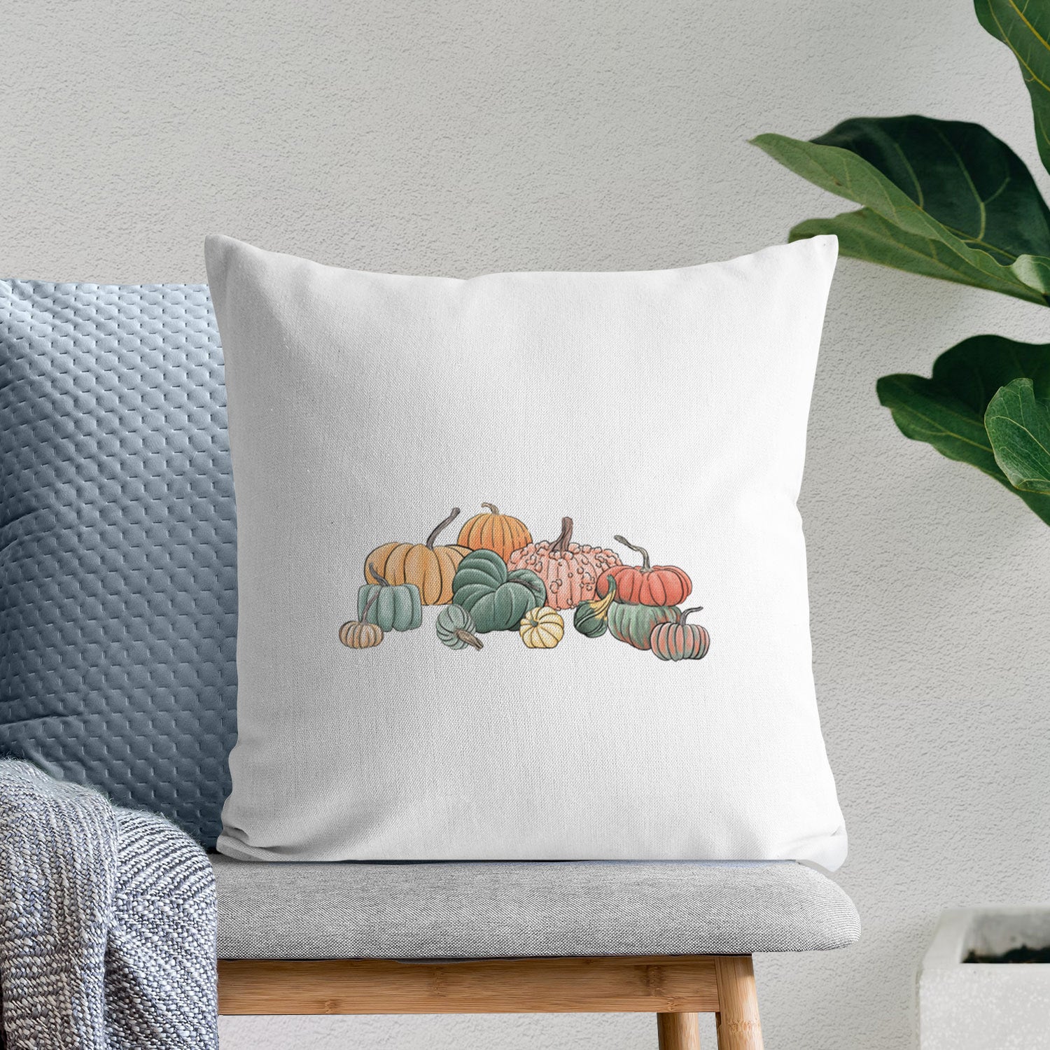 White pillow cover printed with a full-color illustration of a pile of pumpkins and gourds in orange, green and white, on a bench beside plants.