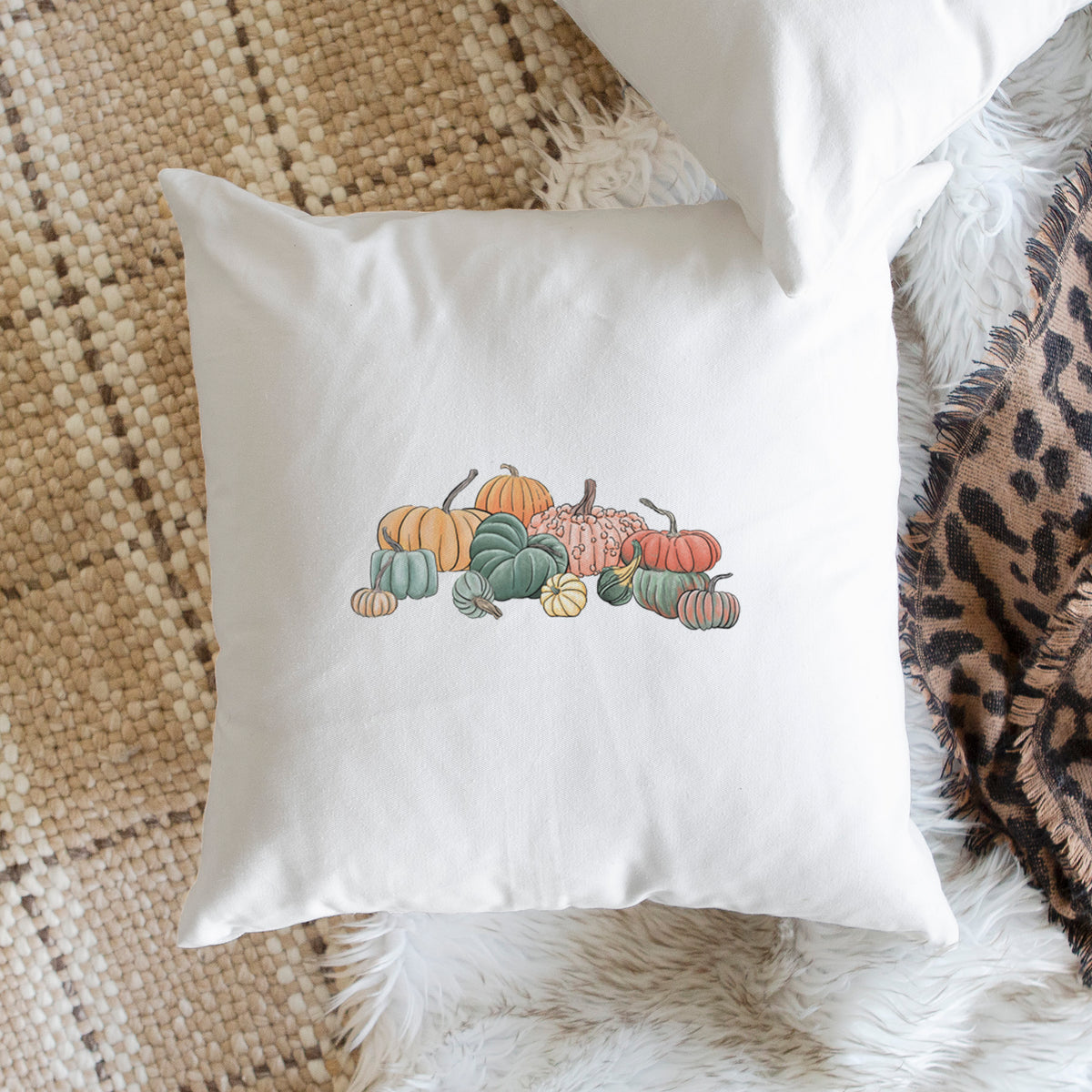 White pillow cover printed with a full-color illustration of a pile of pumpkins and gourds in orange, green and white, on a woven chair.