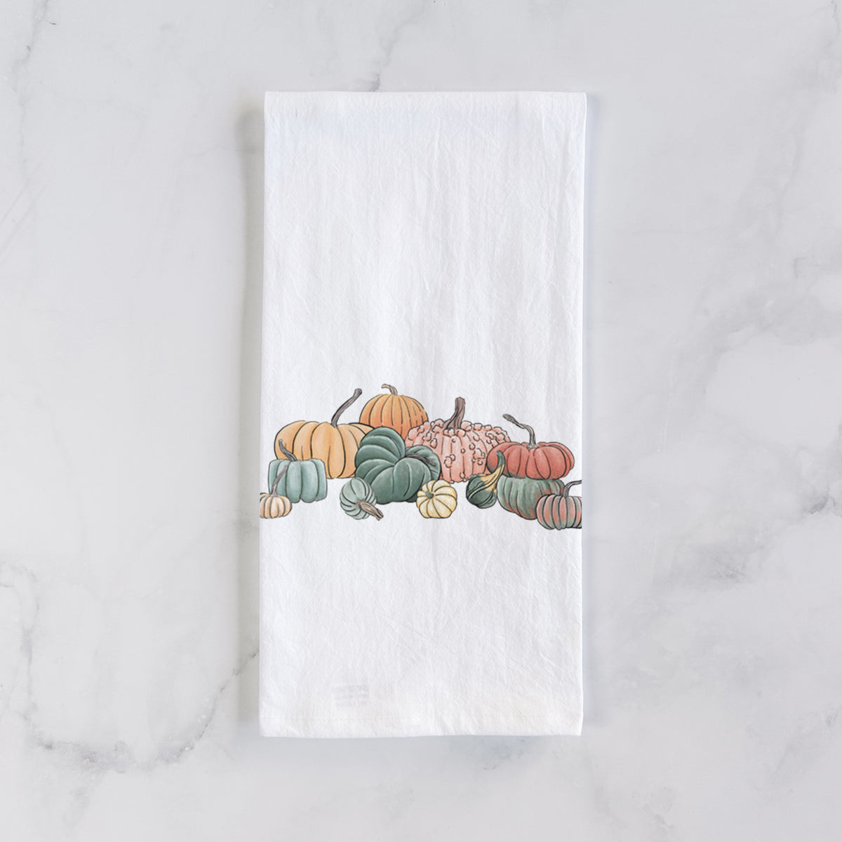 White cotton tea towel laid flat on marble, printed with a full-color illustration of a pile of pumpkins and gourds in orange, green and white.