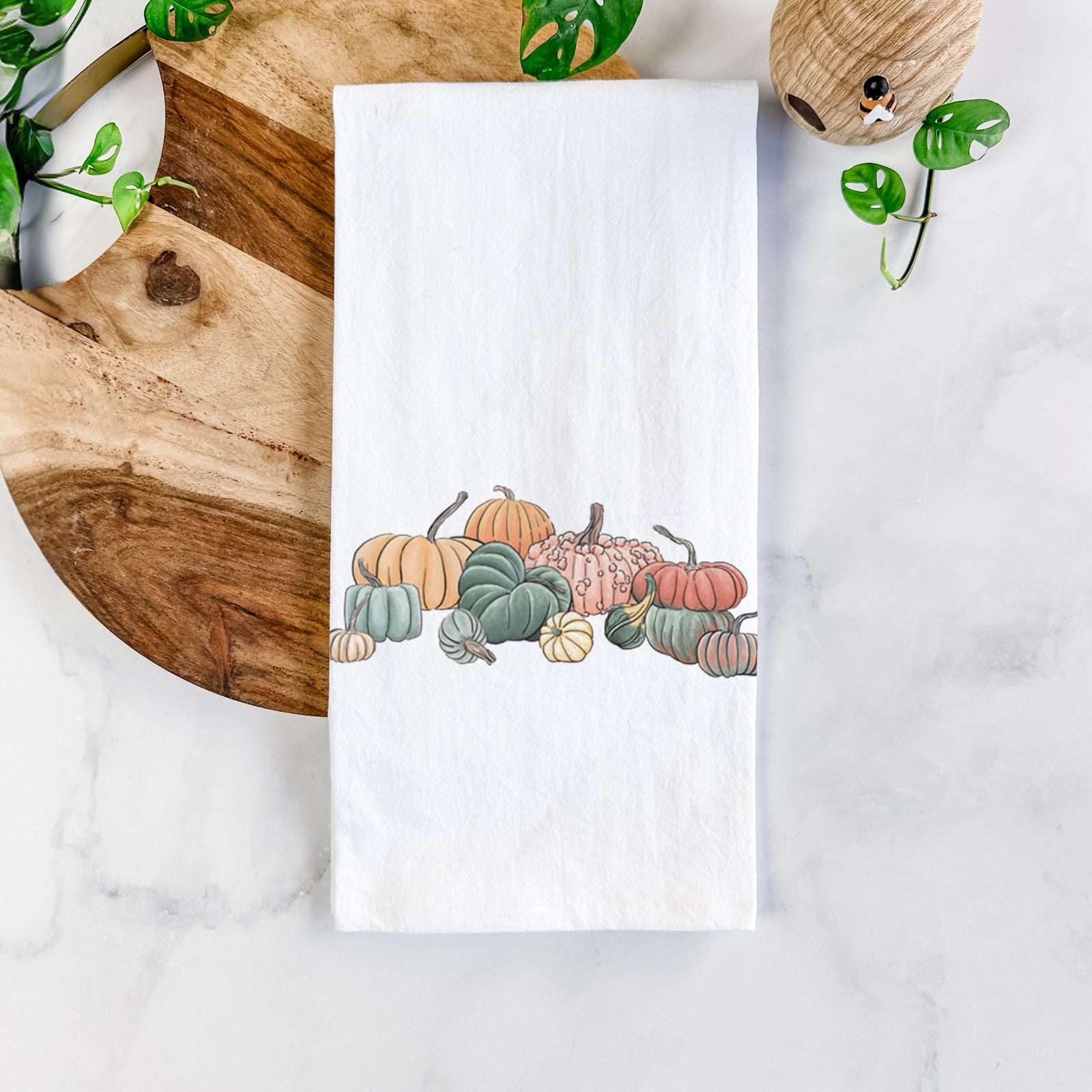 White cotton tea towel printed with a full-color illustration of a pile of pumpkins and gourds in orange, green and white, on a marble counter beside a round wooden cutting board and greenery.