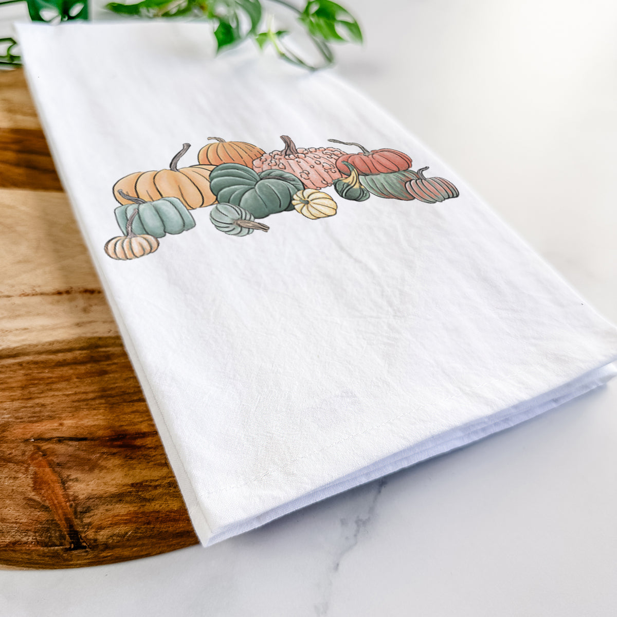Close-up of a folded white tea towel on a wooden board, showing a full-color illustration of a pile of pumpkins and gourds in orange, green and white.