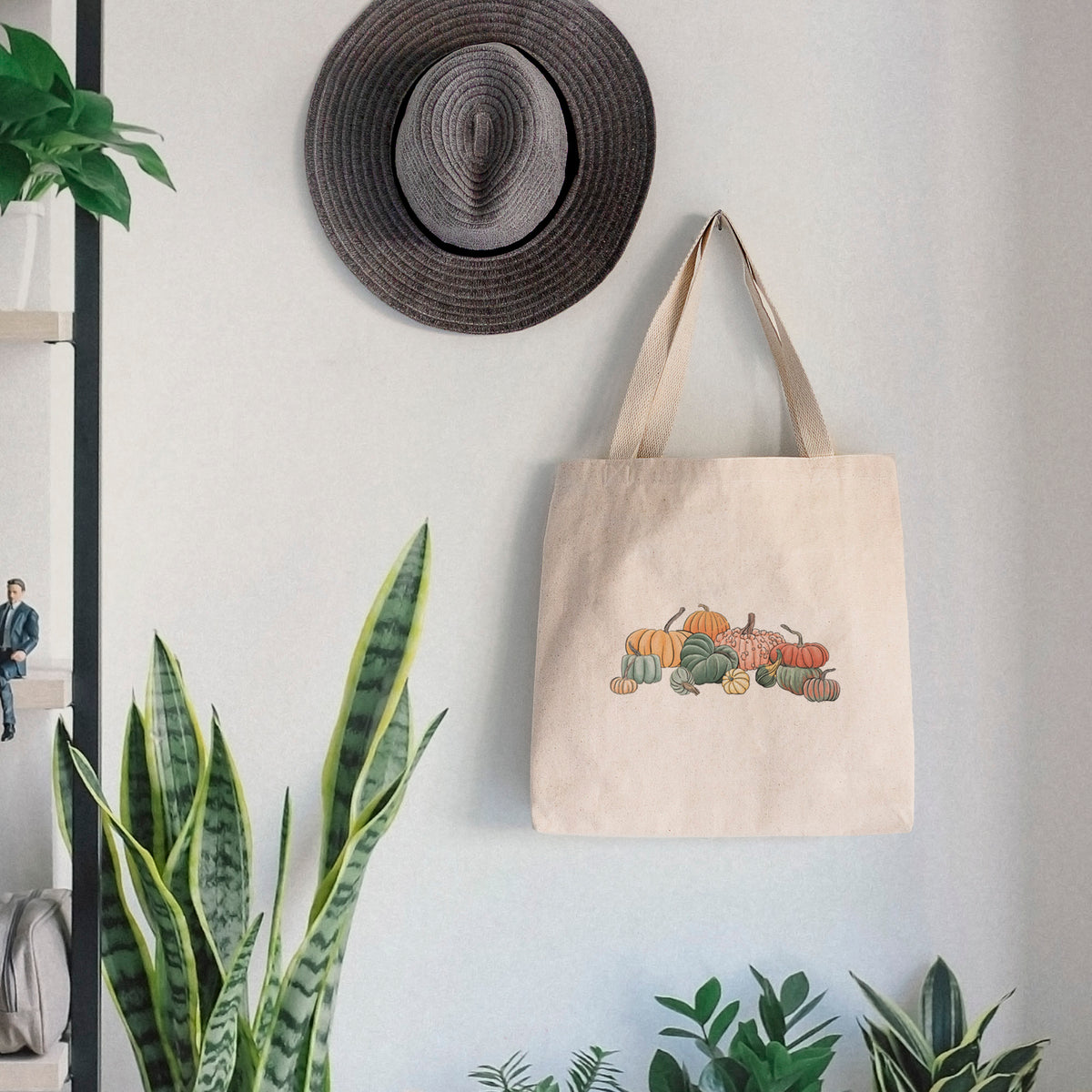 Canvas tote bag printed with a full-color illustration of a pile of pumpkins and gourds in orange, green and white, hanging on a wall hook below a hat, next to potted plants.