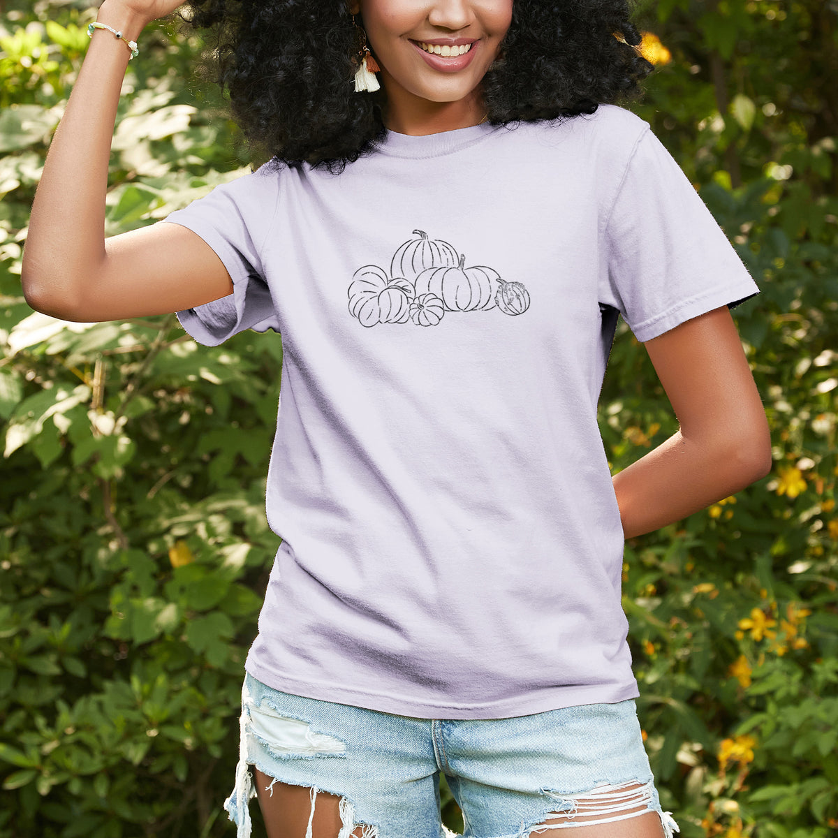Woman wearing a Comfort Colors t-shirt printed with a black ink line drawing of a pile of pumpkins, outdoors.
