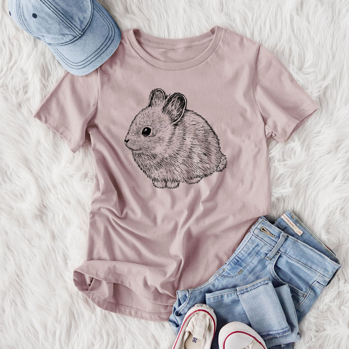 Women's relaxed fit cotton t-shirt printed with a detailed black ink illustration of a round, fluffy pygmy rabbit, styled flat on a white faux fur rug with jeans.