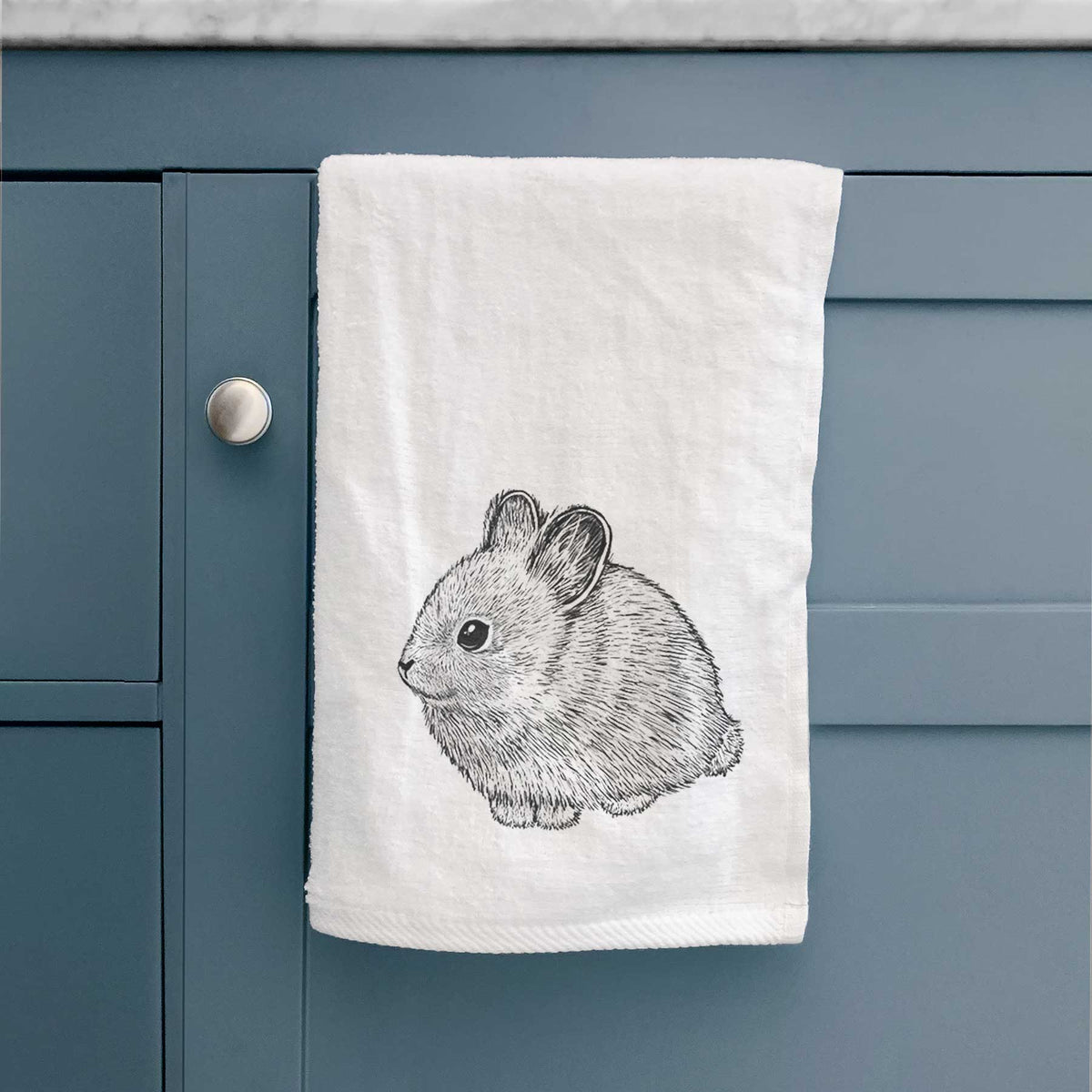 White hand towel printed with a detailed black ink illustration of a round, fluffy pygmy rabbit, hanging on a cabinet door.