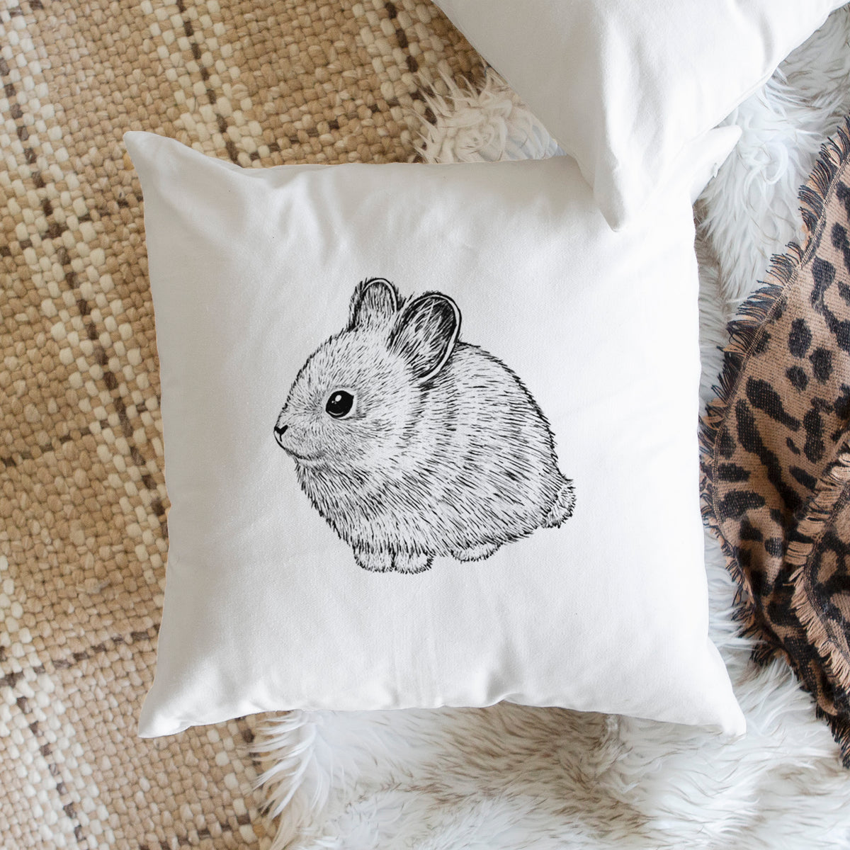 White pillow cover printed with a detailed black ink illustration of a round, fluffy pygmy rabbit, on a woven chair.
