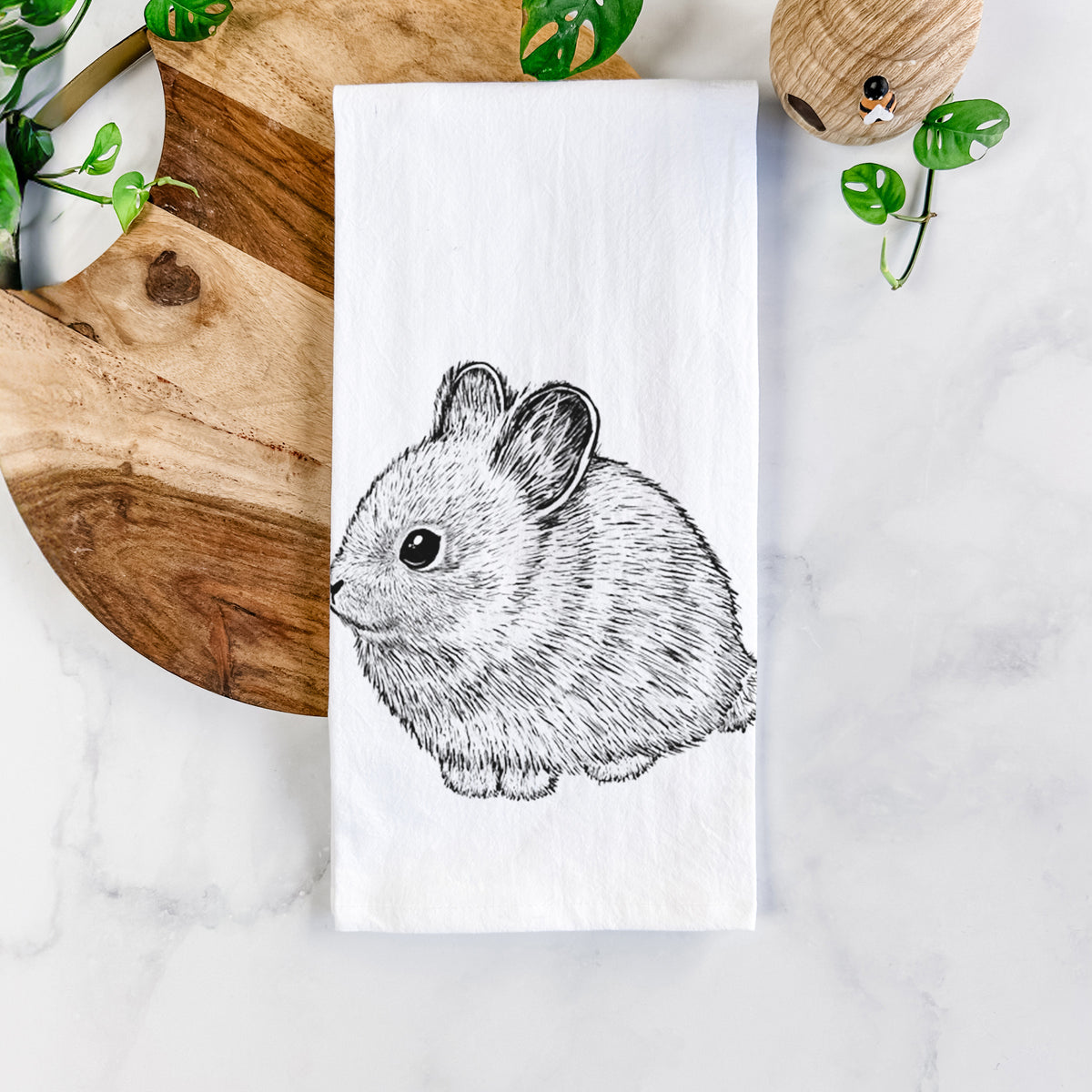 White cotton tea towel printed with a detailed black ink illustration of a round, fluffy pygmy rabbit, on a marble counter beside a round wooden cutting board and greenery.