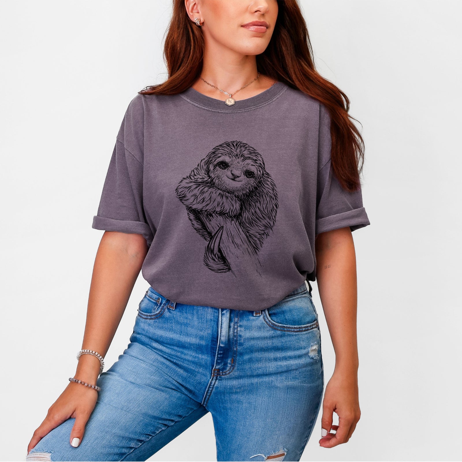 Blue jean Comfort Colors t-shirt laid flat, printed with a detailed black ink illustration of a smiling pygmy three-toed sloth hanging from a branch.