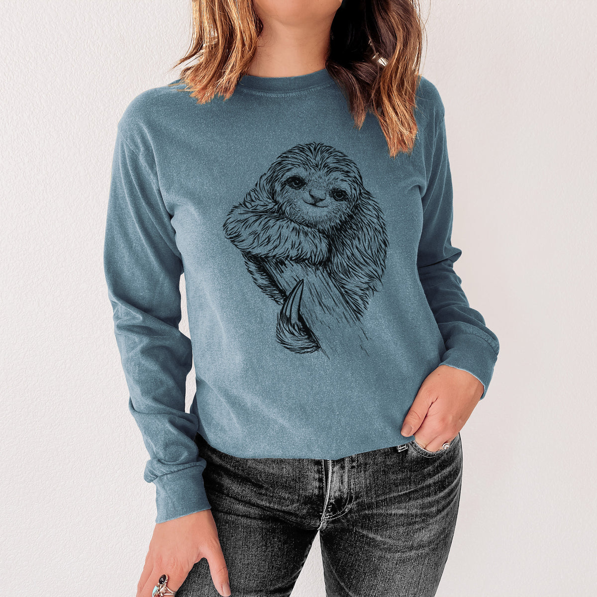 Steel blue Comfort Colors long sleeve shirt printed with a detailed black ink illustration of a smiling pygmy three-toed sloth hanging from a branch, shown in a lifestyle photo.