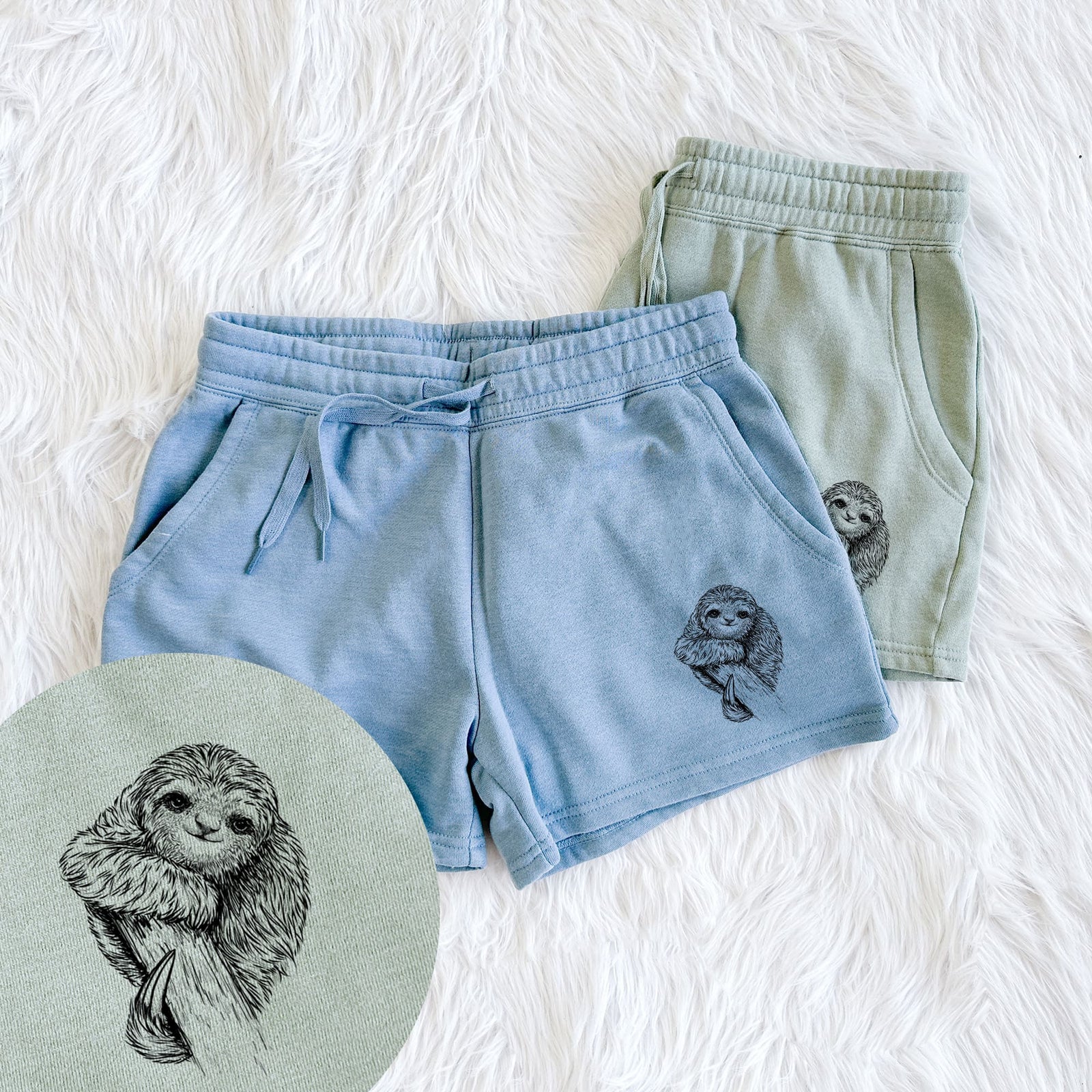 Two pairs of Cali Wave shorts in different colors printed with a detailed black ink illustration of a smiling pygmy three-toed sloth hanging from a branch, with a close-up of the print.