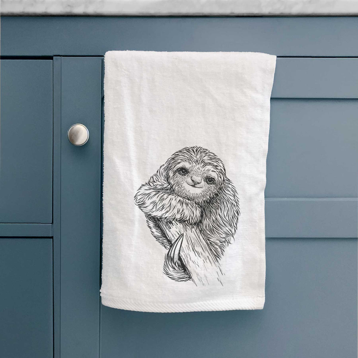 White hand towel printed with a detailed black ink illustration of a smiling pygmy three-toed sloth hanging from a branch, hanging on a cabinet door.