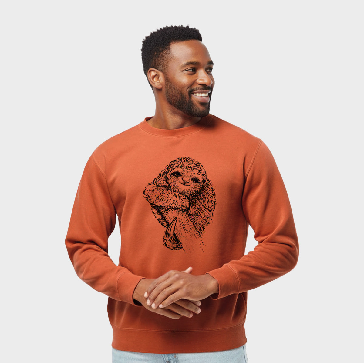 Model wearing an amber orange pigment dyed crewneck sweatshirt printed with a detailed black ink illustration of a smiling pygmy three-toed sloth hanging from a branch.
