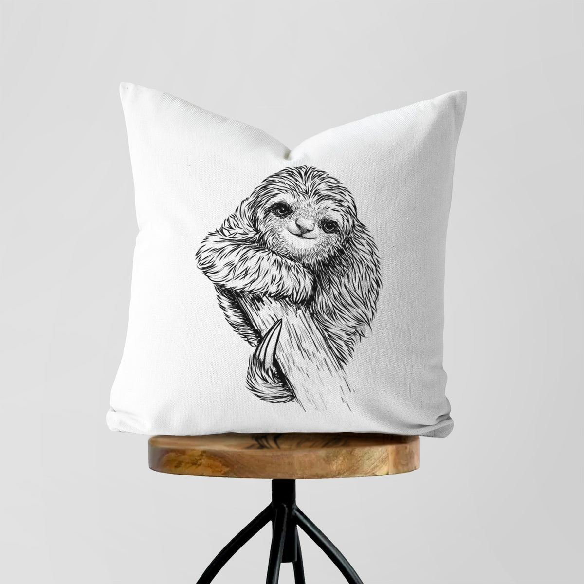 White pillow cover printed with a detailed black ink illustration of a smiling pygmy three-toed sloth hanging from a branch, on a wooden stool.