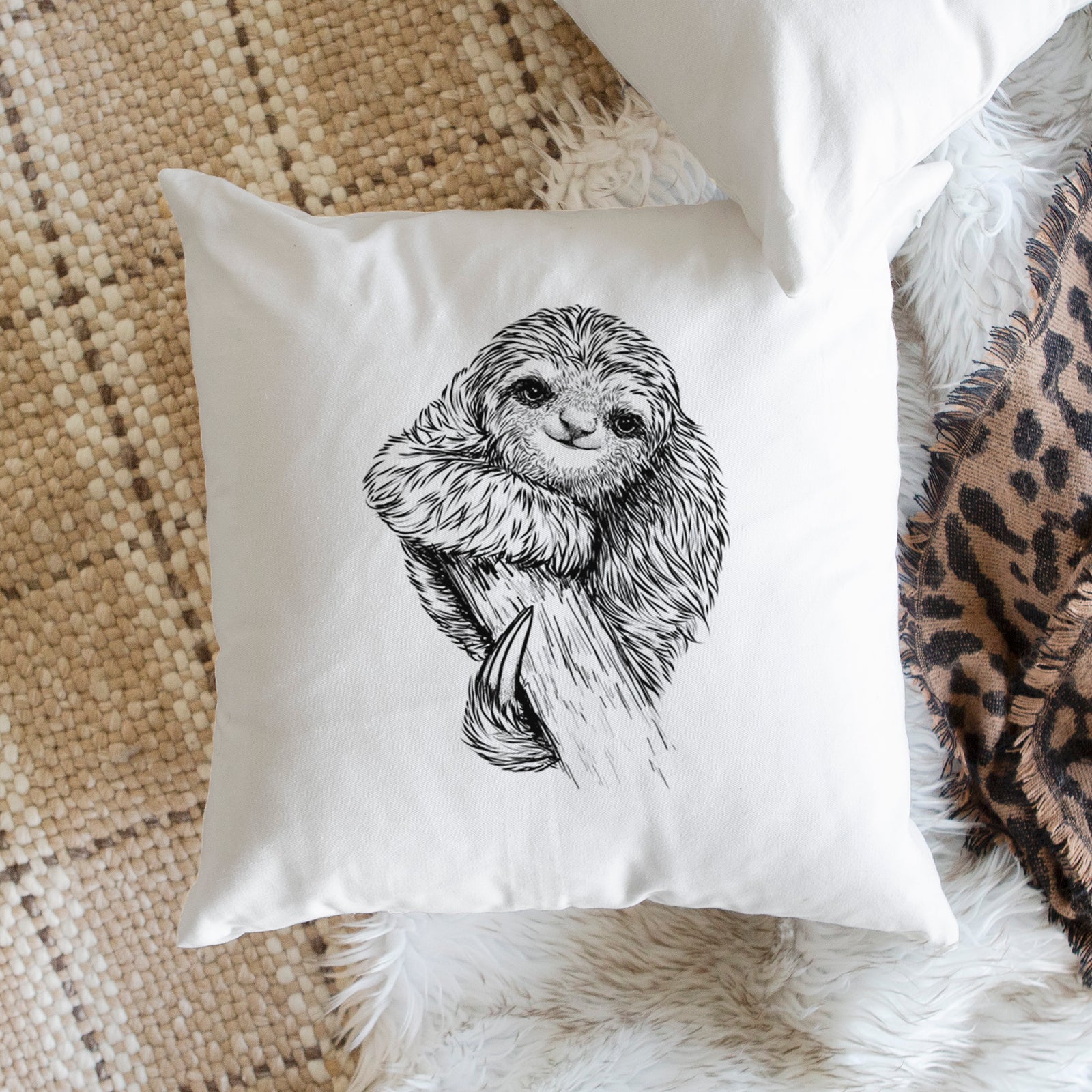 White pillow cover printed with a detailed black ink illustration of a smiling pygmy three-toed sloth hanging from a branch, on a bench beside plants.