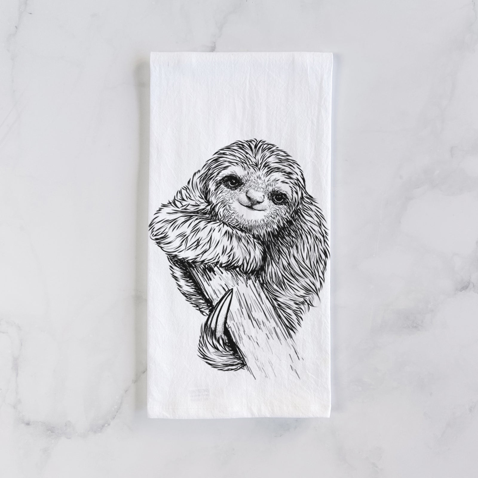 White cotton tea towel printed with a detailed black ink illustration of a smiling pygmy three-toed sloth hanging from a branch, on a marble counter beside a round wooden cutting board and greenery.