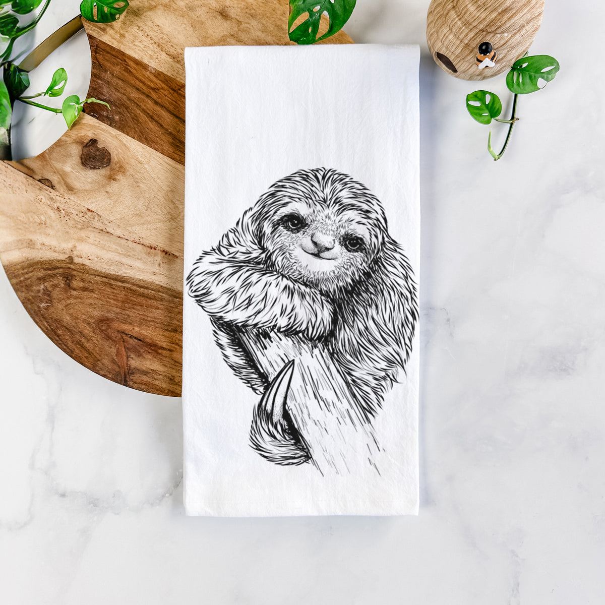 White cotton tea towel printed with a detailed black ink illustration of a smiling pygmy three-toed sloth hanging from a branch, on a marble counter beside a round wooden cutting board and greenery.