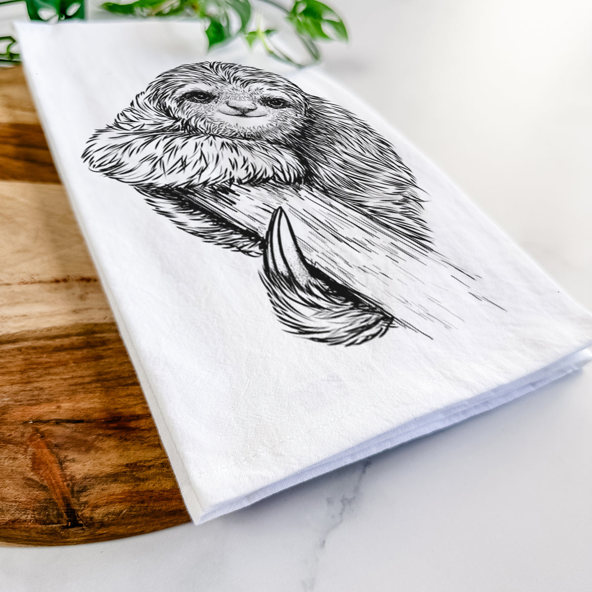 Close-up of a folded white tea towel on a wooden board, showing a detailed black ink illustration of a smiling pygmy three-toed sloth hanging from a branch.