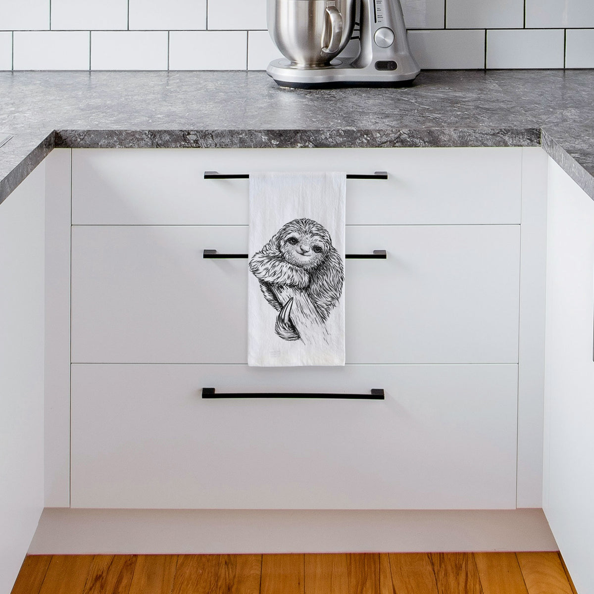 White tea towel printed with a detailed black ink illustration of a smiling pygmy three-toed sloth hanging from a branch, hanging from a drawer handle in a bright white kitchen.