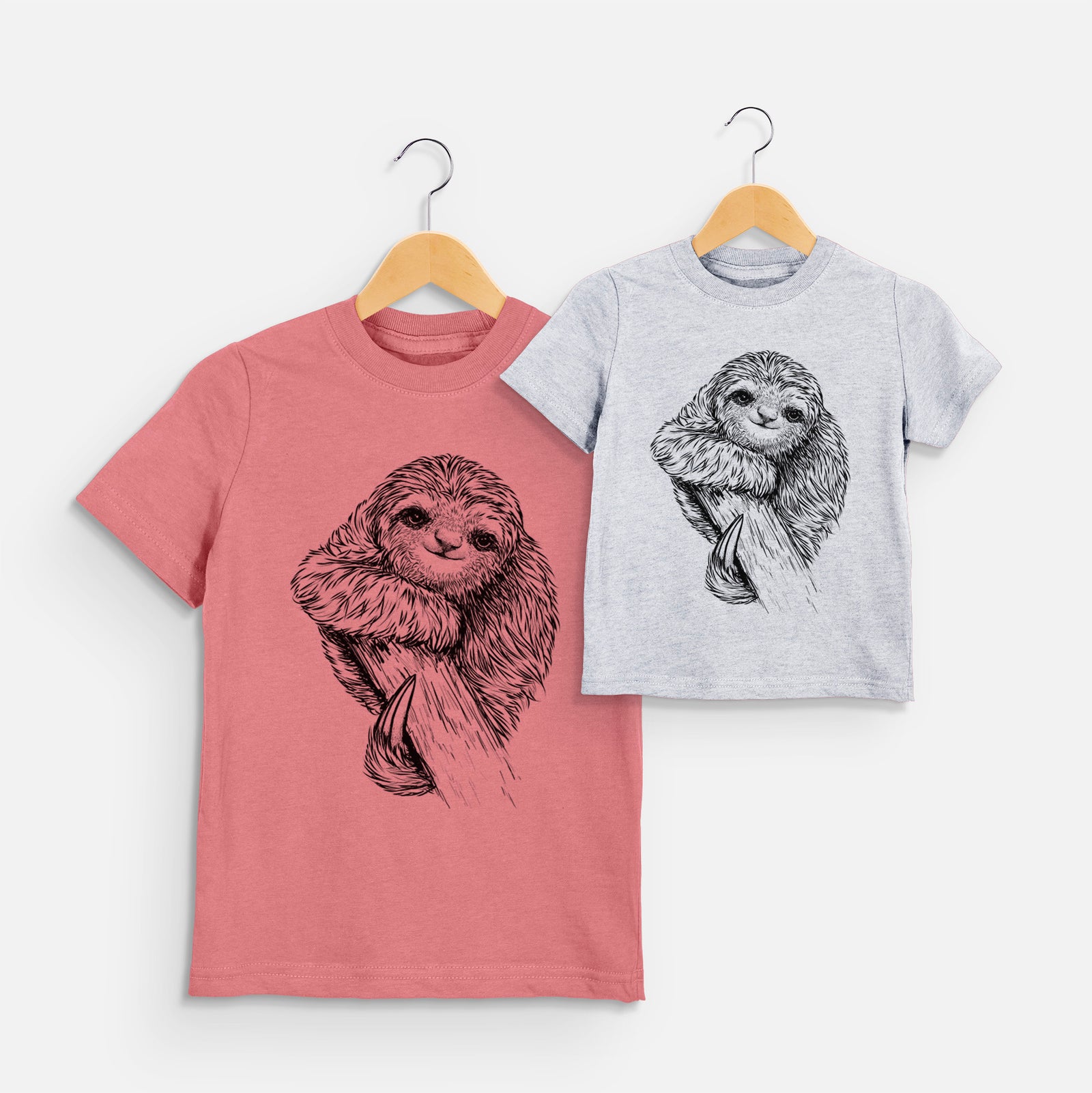 Two youth t-shirts in different colors on hangers, printed with a detailed black ink illustration of a smiling pygmy three-toed sloth hanging from a branch.