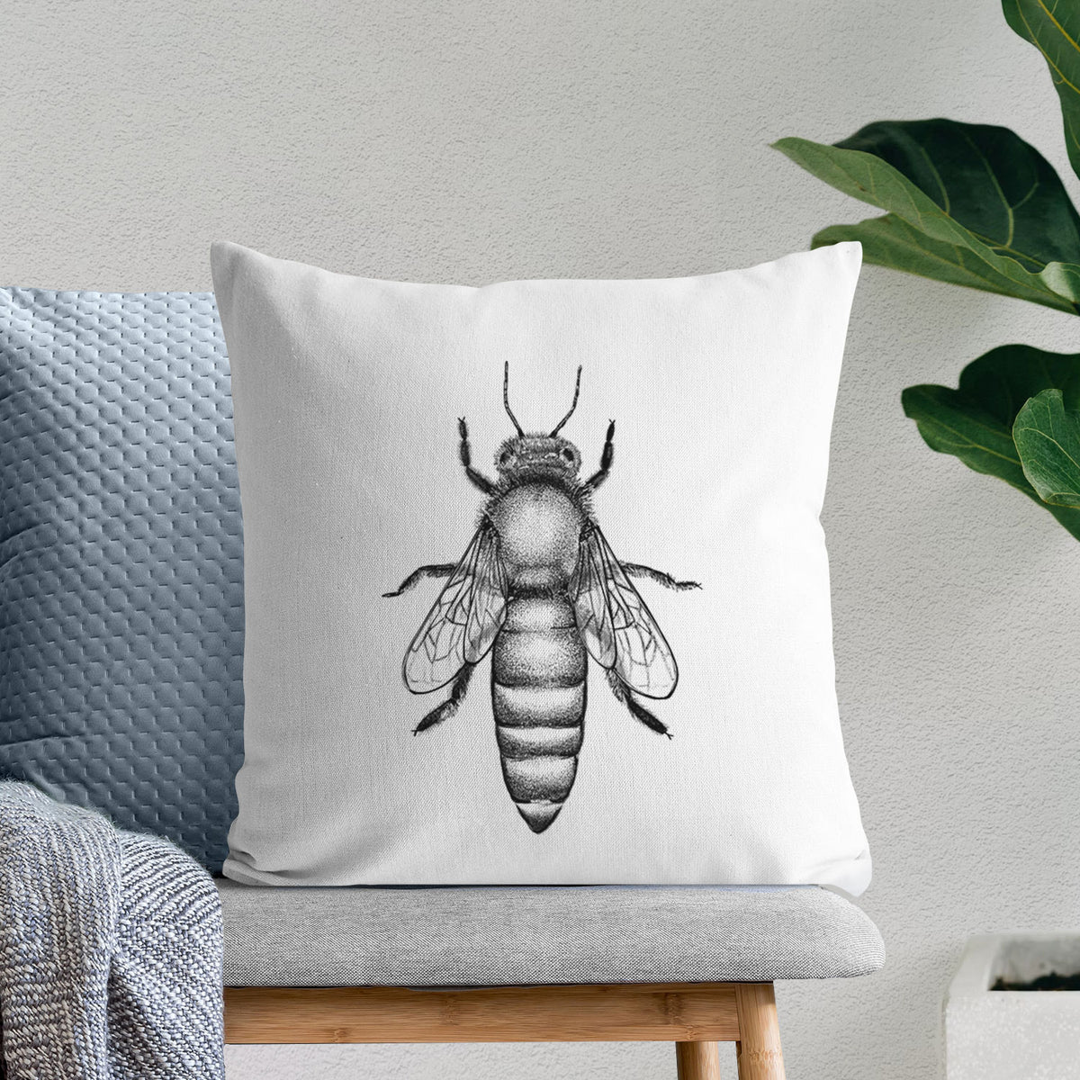 White pillow cover printed with a detailed black ink illustration of a queen honey bee, seen from above, on a bench beside plants.