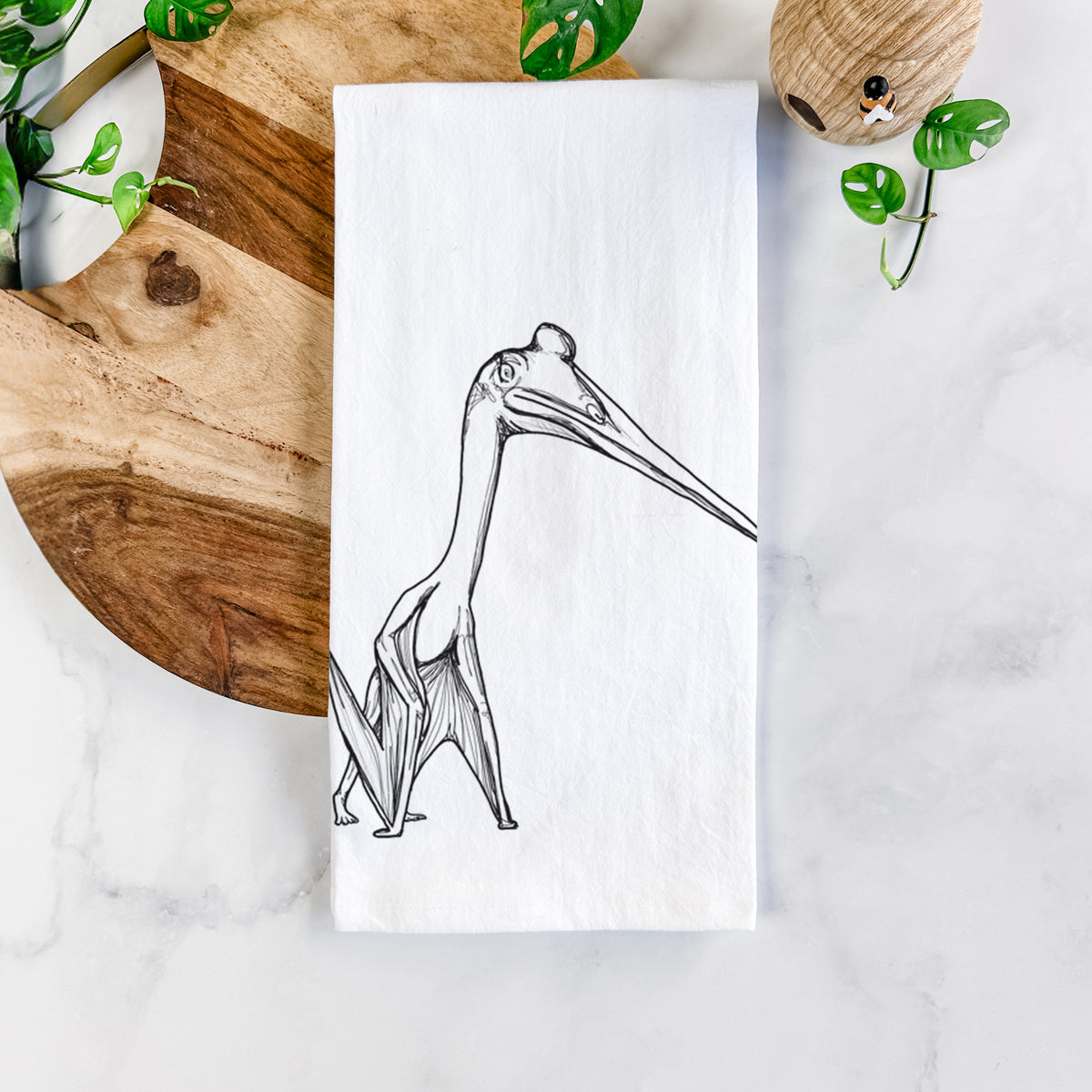White cotton tea towel printed with a black ink illustration of a Quetzalcoatlus pterosaur standing on all fours, on a marble counter beside a round wooden cutting board and greenery.