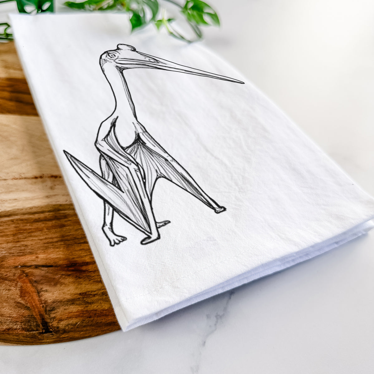 Close-up of a folded white tea towel on a wooden board, showing a black ink illustration of a Quetzalcoatlus pterosaur standing on all fours.