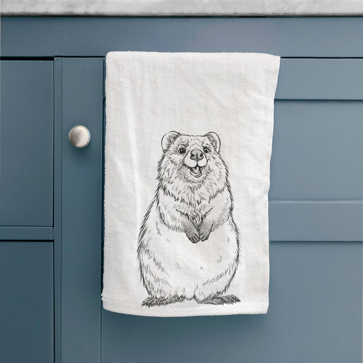 White hand towel printed with a black ink illustration of a smiling quokka sitting upright, hanging on a cabinet door.
