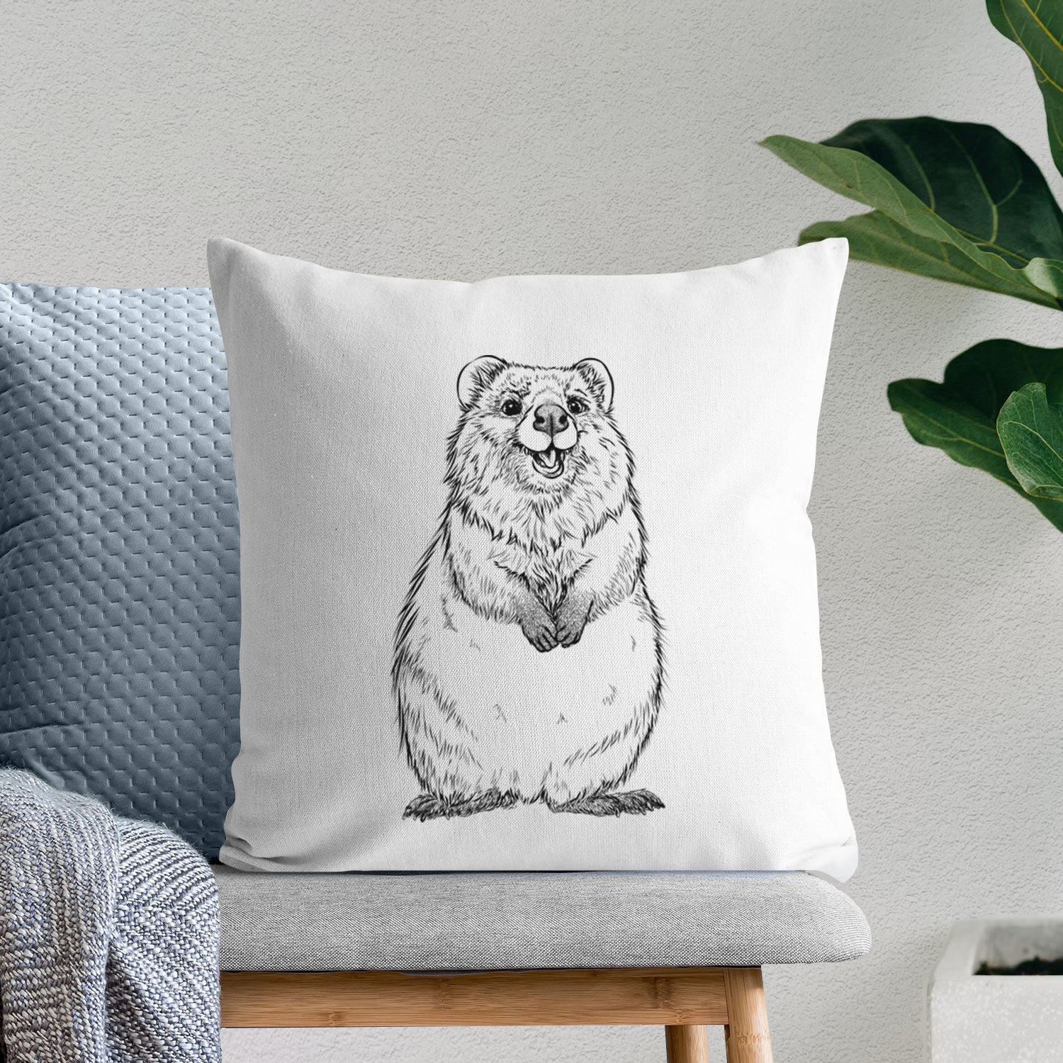 White pillow cover printed with a black ink illustration of a smiling quokka sitting upright, on a bench beside plants.