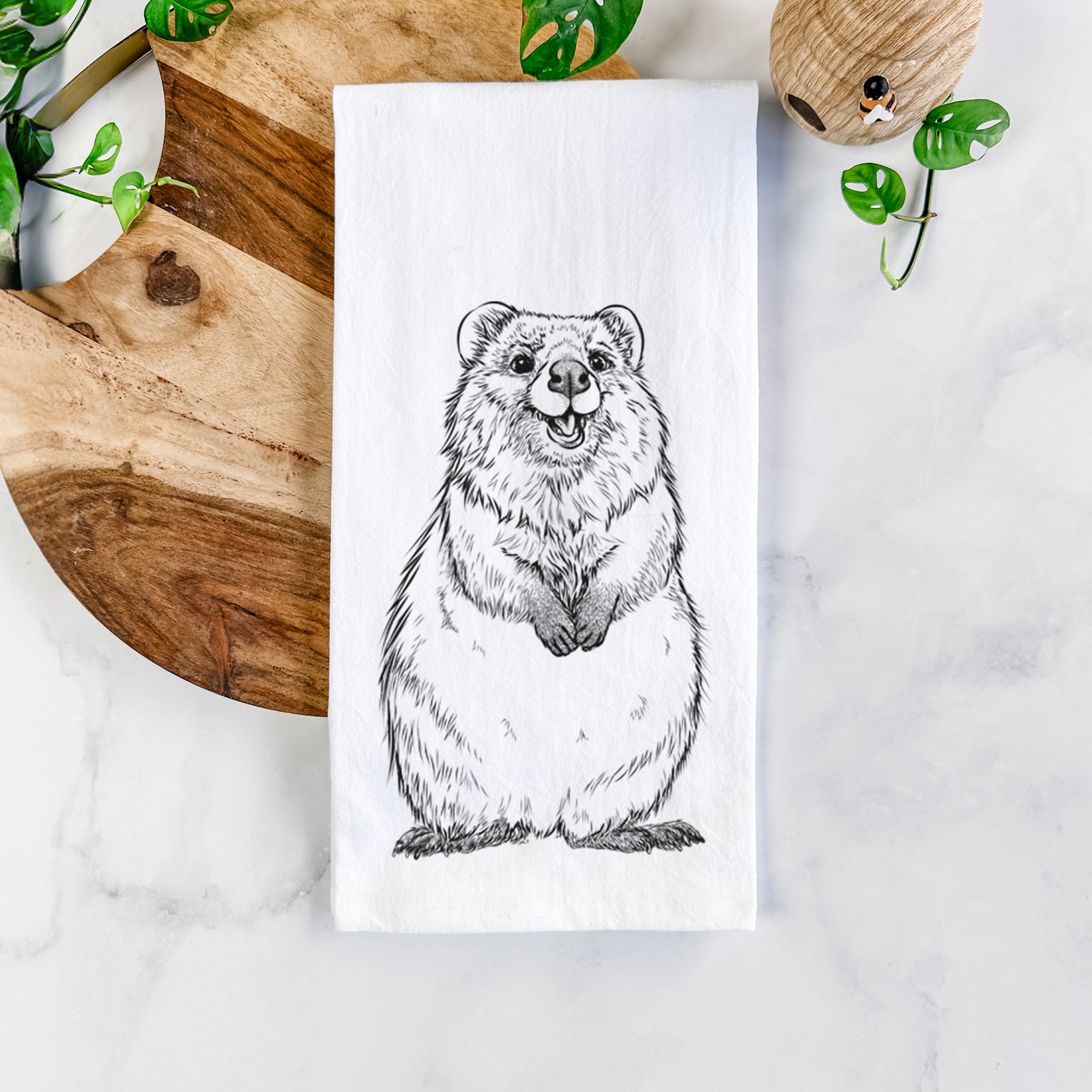 White cotton tea towel printed with a black ink illustration of a smiling quokka sitting upright, on a marble counter beside a round wooden cutting board and greenery.