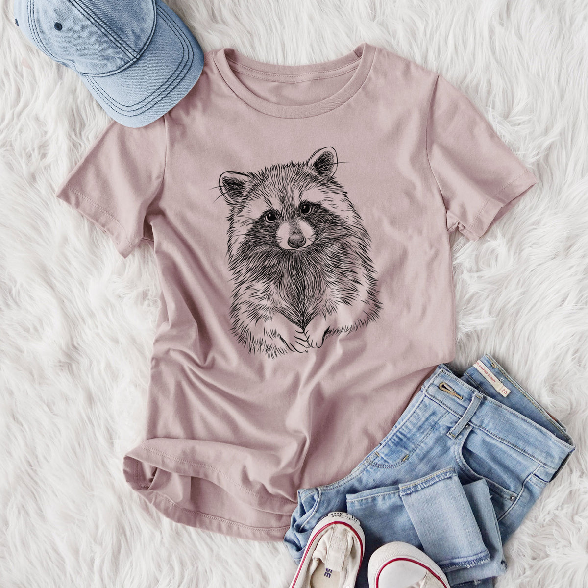 Women's relaxed fit cotton t-shirt printed with a detailed black ink portrait of a raccoon with its front paws resting in front of it, styled flat on a white faux fur rug with jeans.