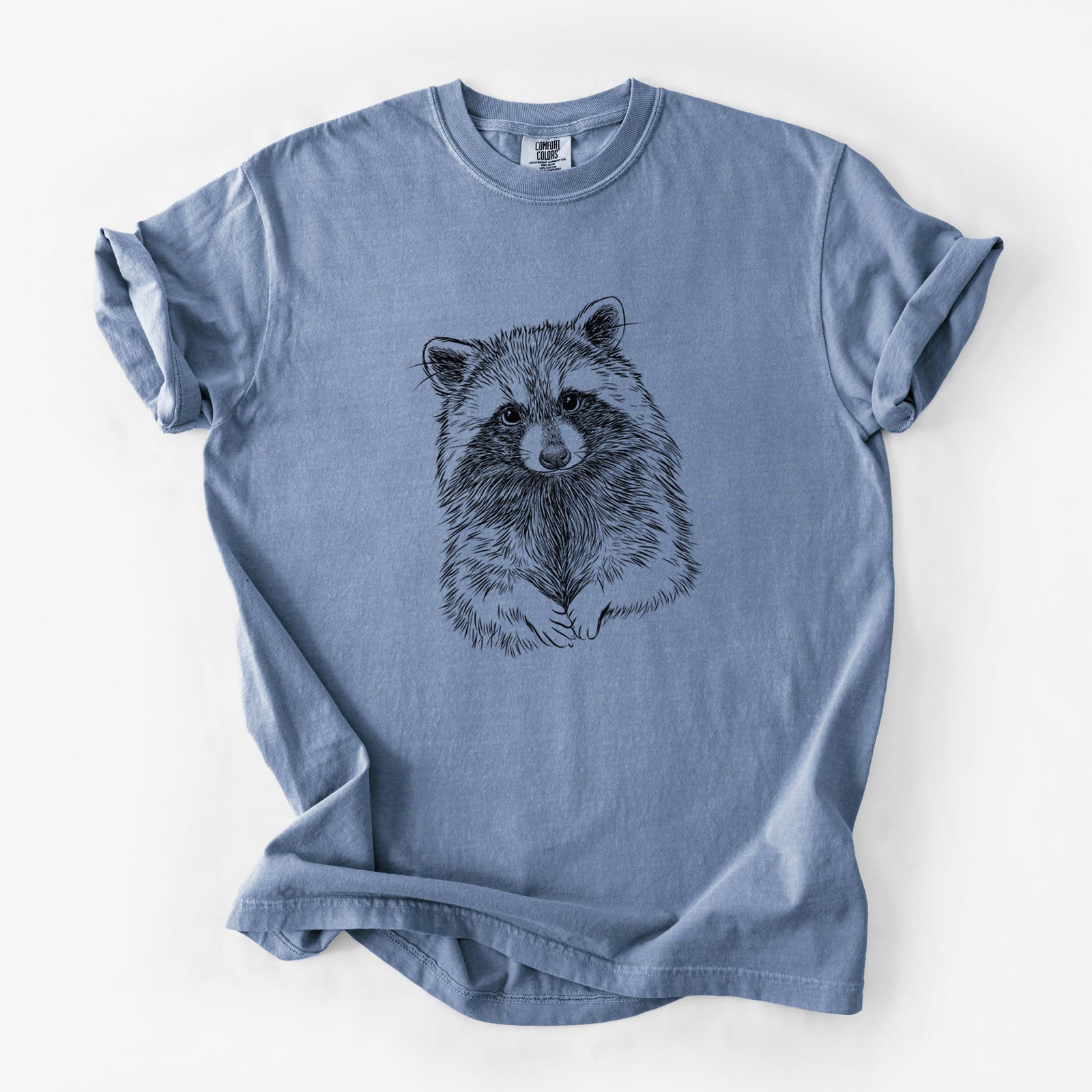 Blue jean Comfort Colors t-shirt laid flat, printed with a detailed black ink portrait of a raccoon with its front paws resting in front of it.