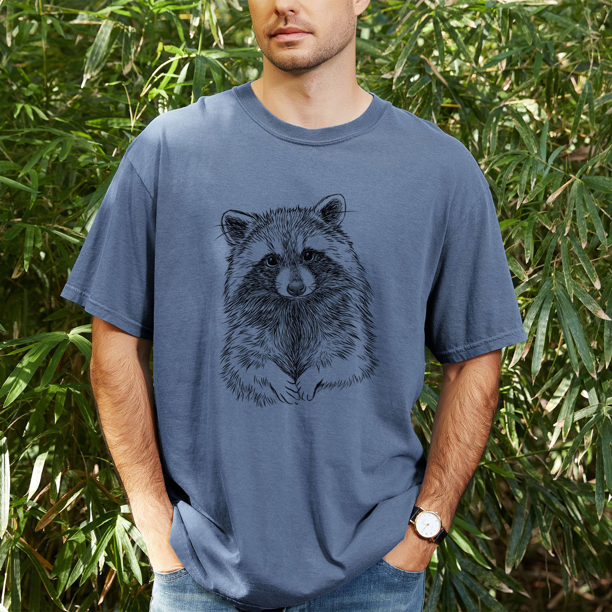 Man wearing a Comfort Colors t-shirt printed with a detailed black ink portrait of a raccoon with its front paws resting in front of it, outdoors.