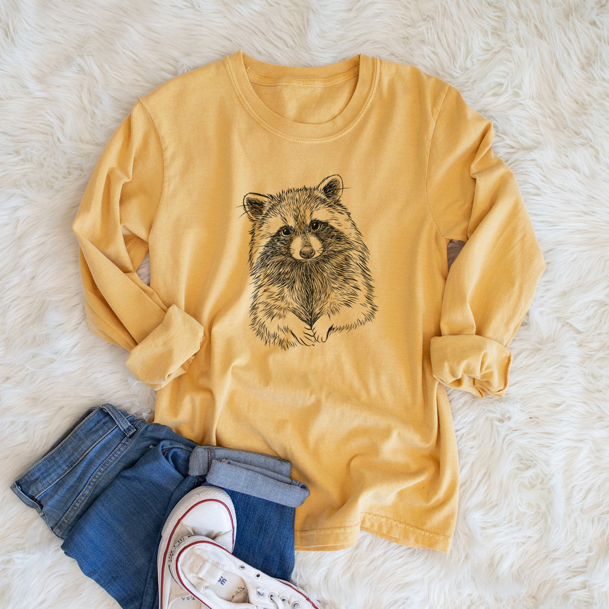 Golden wheat yellow Comfort Colors long sleeve shirt printed with a detailed black ink portrait of a raccoon with its front paws resting in front of it, shown in a lifestyle photo.