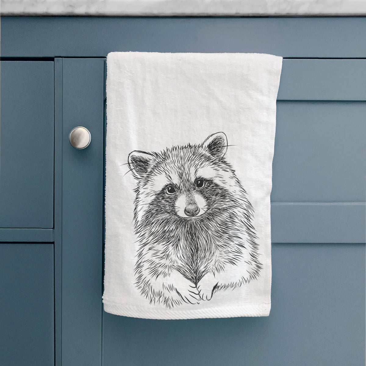 White hand towel printed with a detailed black ink portrait of a raccoon with its front paws resting in front of it, hanging on a cabinet door.